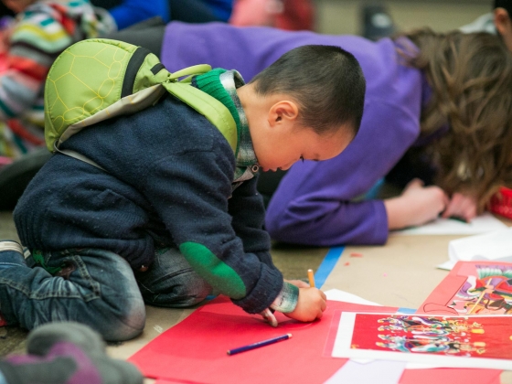 Young visitor with turtle shell backpack kneels on floor drawing on red paper Young visitor with turtle shell backpack kneels on floor drawing on red paper