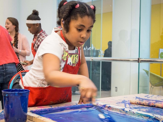Young visitor in red apron dipping brush into tray of blue paint