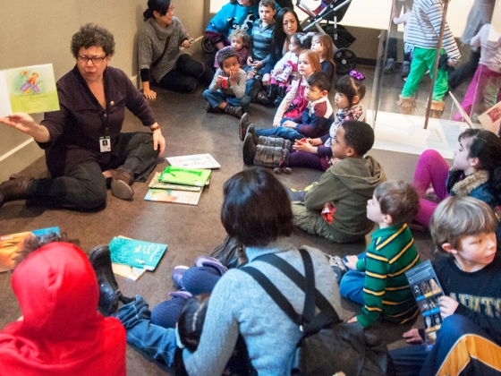 Children sitting on gallery floor around storyteller holding up book Children sitting on gallery floor around storyteller holding up book