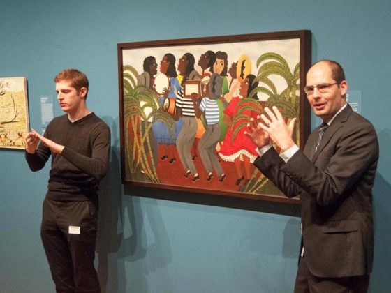 Curator standing next to painting, talking animatedly, with American Sign Language interpreter on opposite side of painting Curator standing next to painting, talking animatedly, with American Sign Language interpreter on opposite side of painting