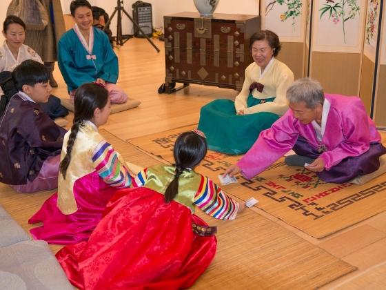 Demonstration of saebae; man in traditional Korean attire reaches forward to place paper in front of two young girls Demonstration of saebae; man in traditional Korean attire reaches forward to place paper in front of two young girls
