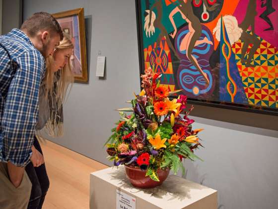 Two visitors crouch down to examine flower arrangement in front of Loïs Mailou Jones' La Baker painting